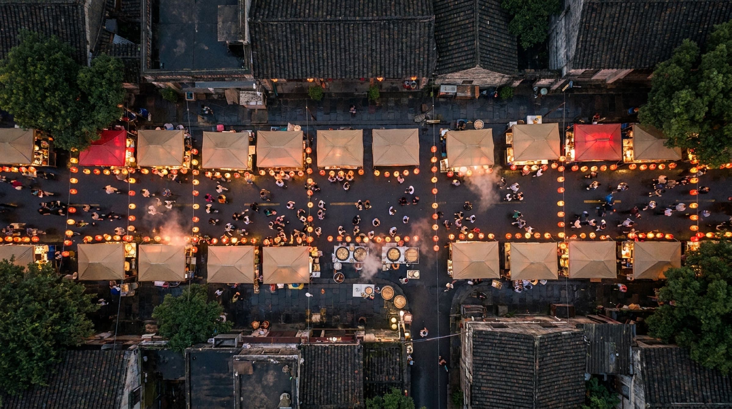 Overhead view of an evening night market street lined with canopied food stalls and rows of glowing orange lanterns, crowds moving between the stalls and steam rising from the cooking.