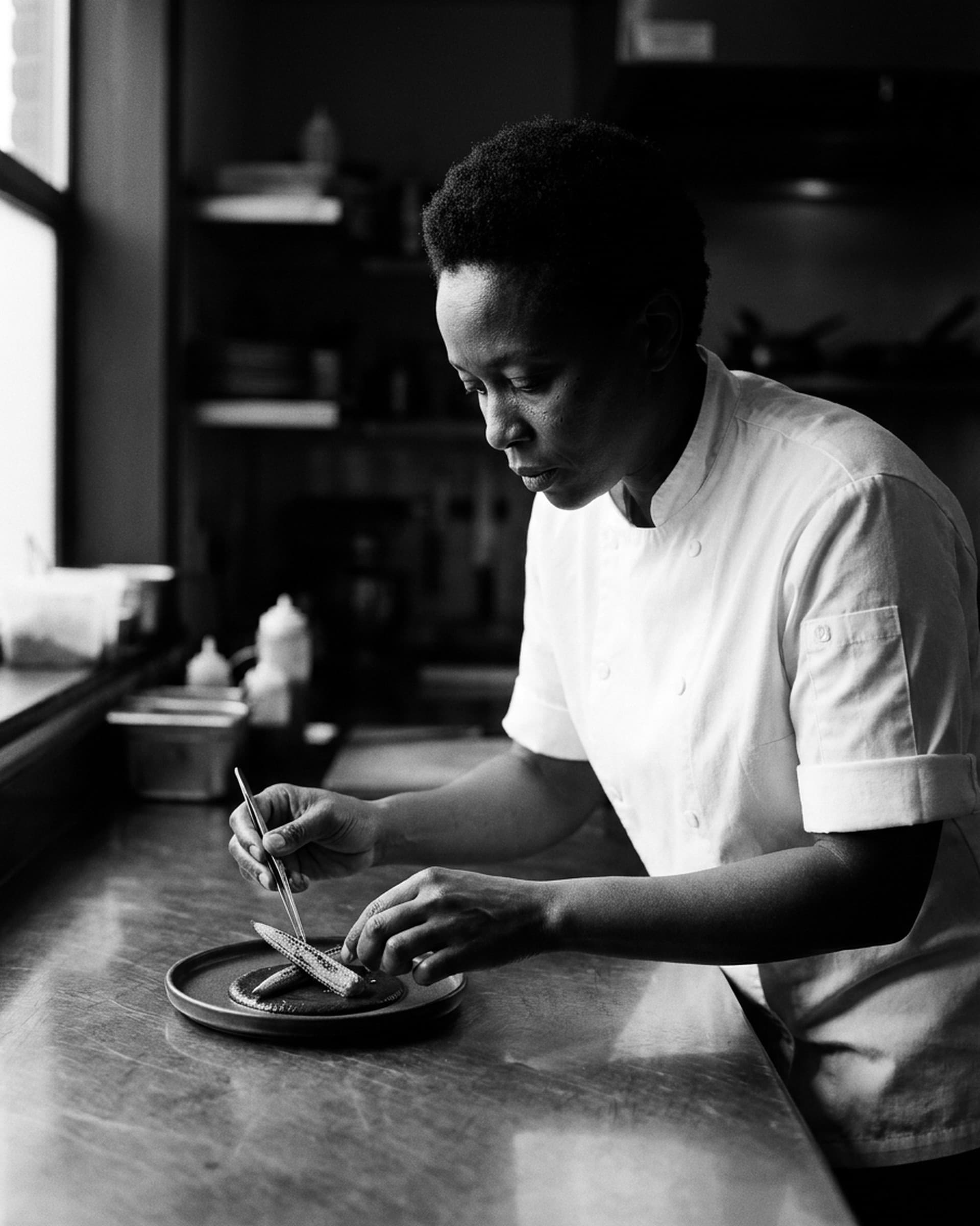 A chef in a white chef coat leans over a steel counter by a window, setting charred baby corn onto a small plate of dark mole with tweezers.