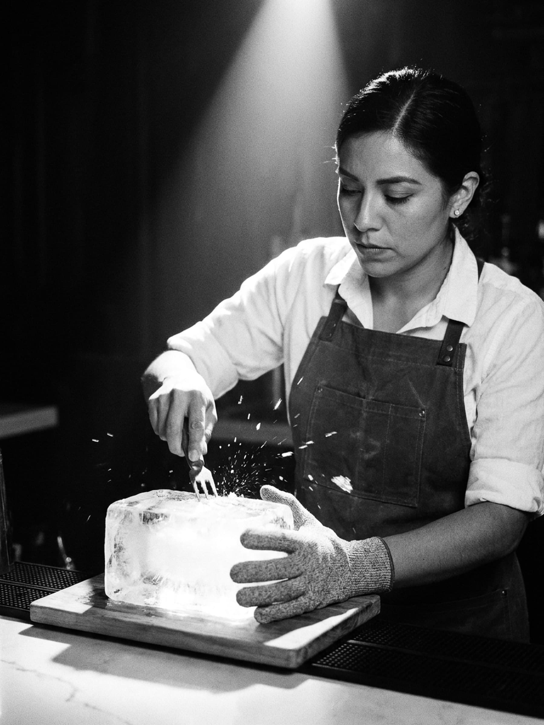 A bartender in a canvas apron and a cut-resistant glove carves a block of clear ice with a three-prong pick, chips of ice flying under a single overhead light.