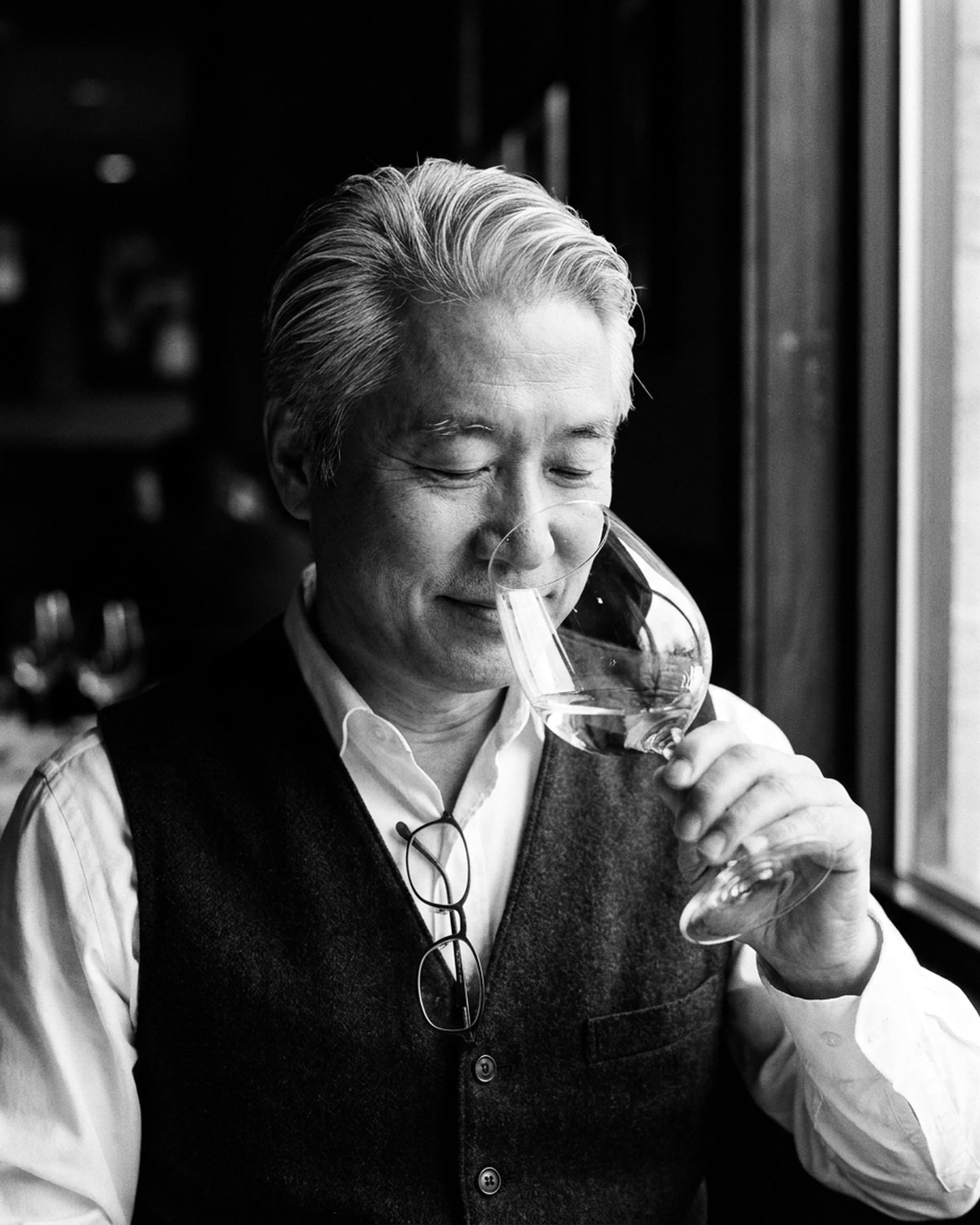 A silver-haired sommelier in a wool vest closes his eyes and smiles slightly as he smells a glass of white wine beside a window.