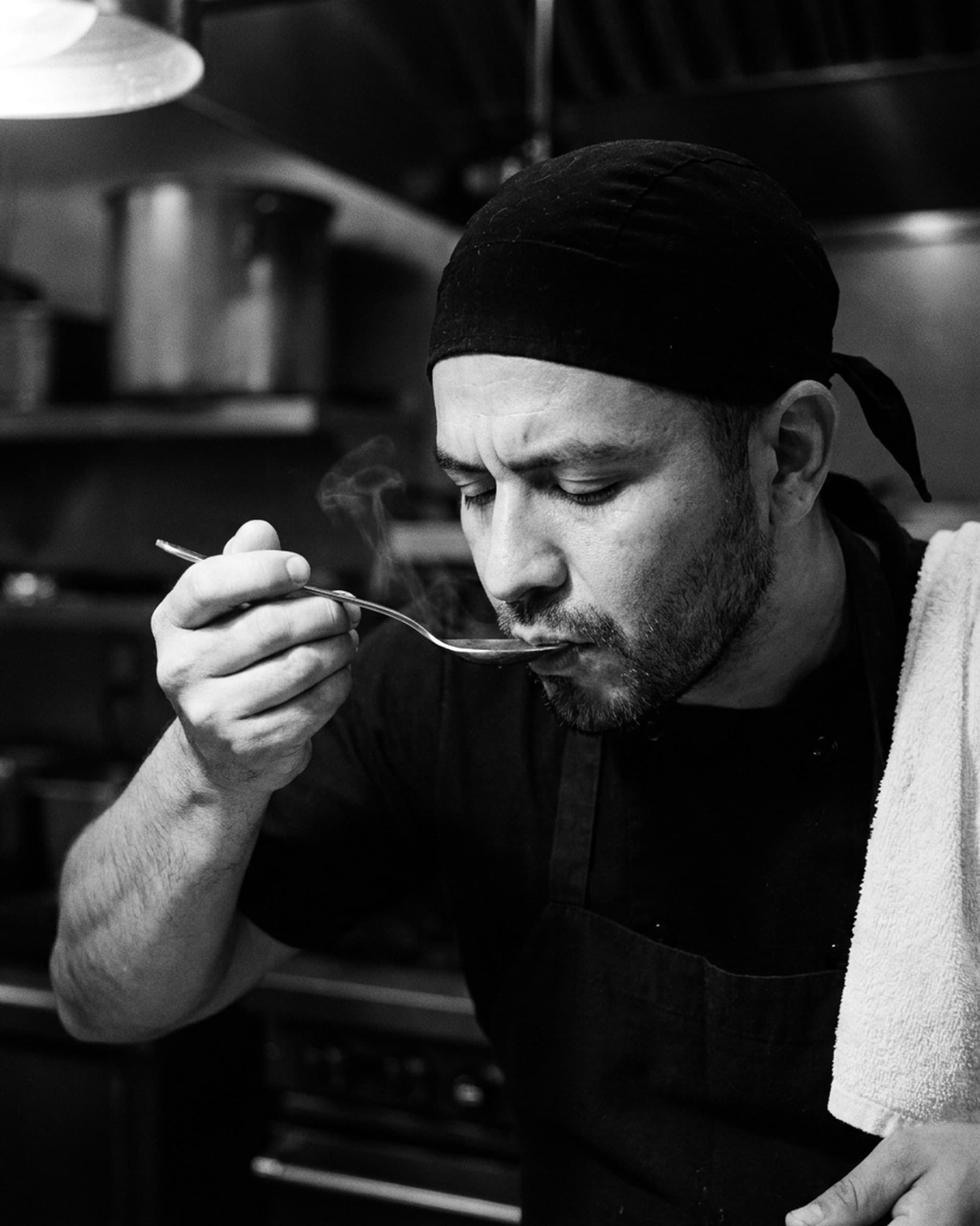A line cook in a black bandana with a towel over his shoulder tastes a steaming spoonful of sauce in a dark kitchen.