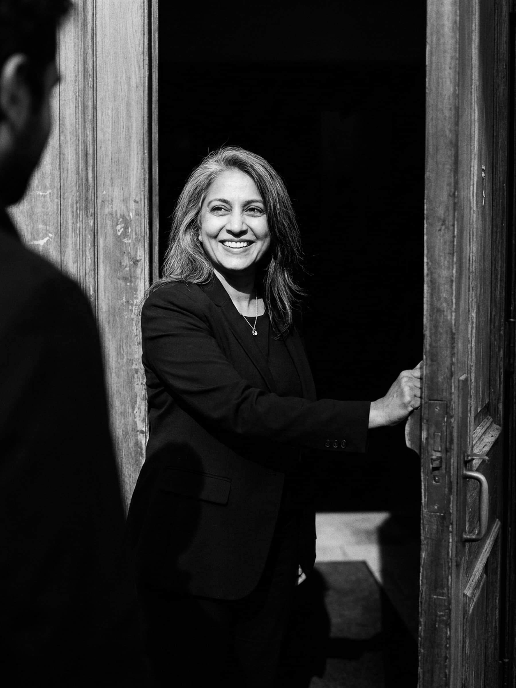 A host in a black blazer pulls open a heavy wooden door and greets an arriving guest with a wide smile.