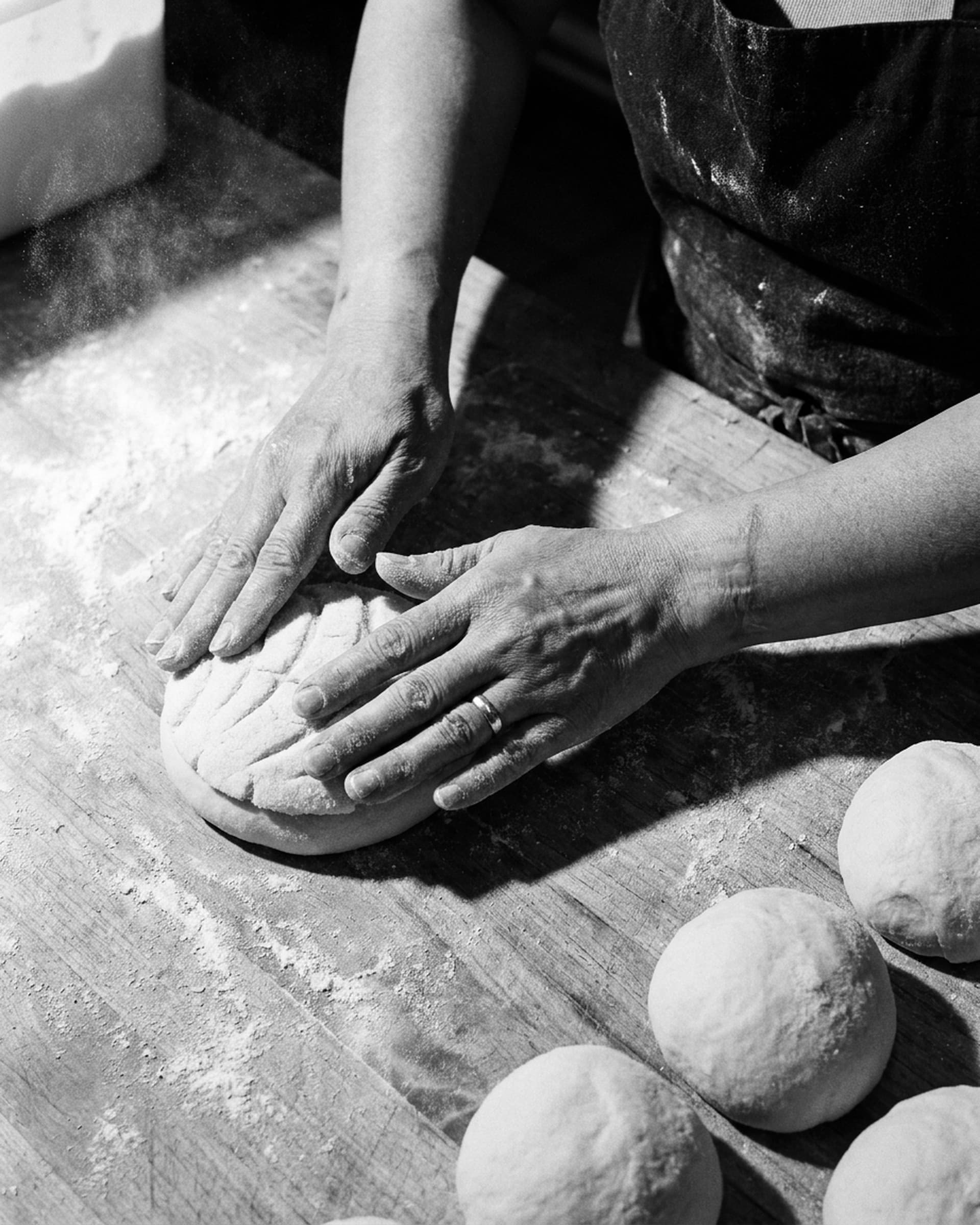 Floured hands press a scored sugar topping onto a round of sweet bread dough on a wooden bench, more dough rounds lined up beside them.