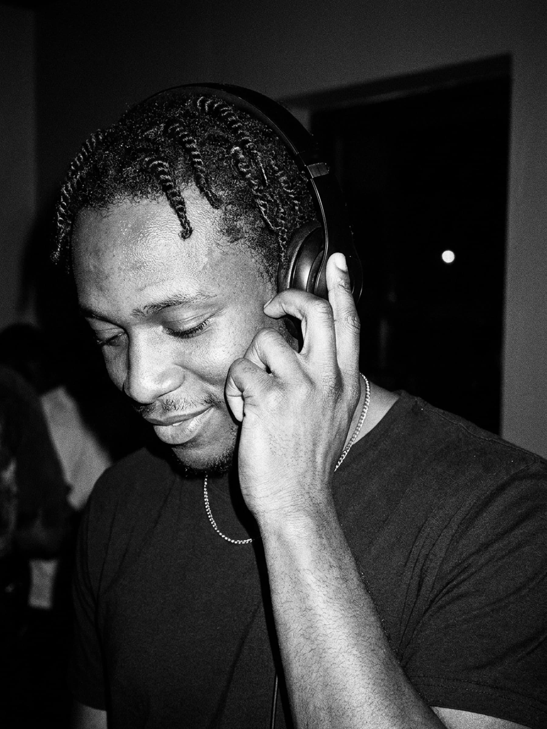 A DJ in a dark tee and a thin chain holds one headphone to his ear, eyes down and smiling, lit by flash in a dark club.