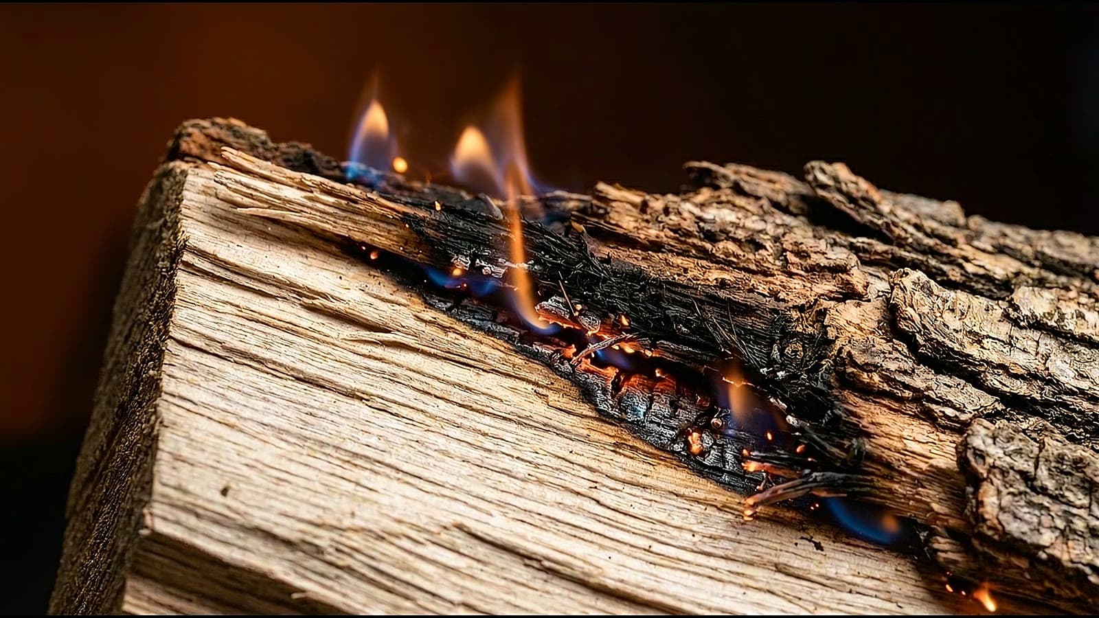 Small flames flicker along the burning seam of an oak log as embers pulse and a spark drifts up.