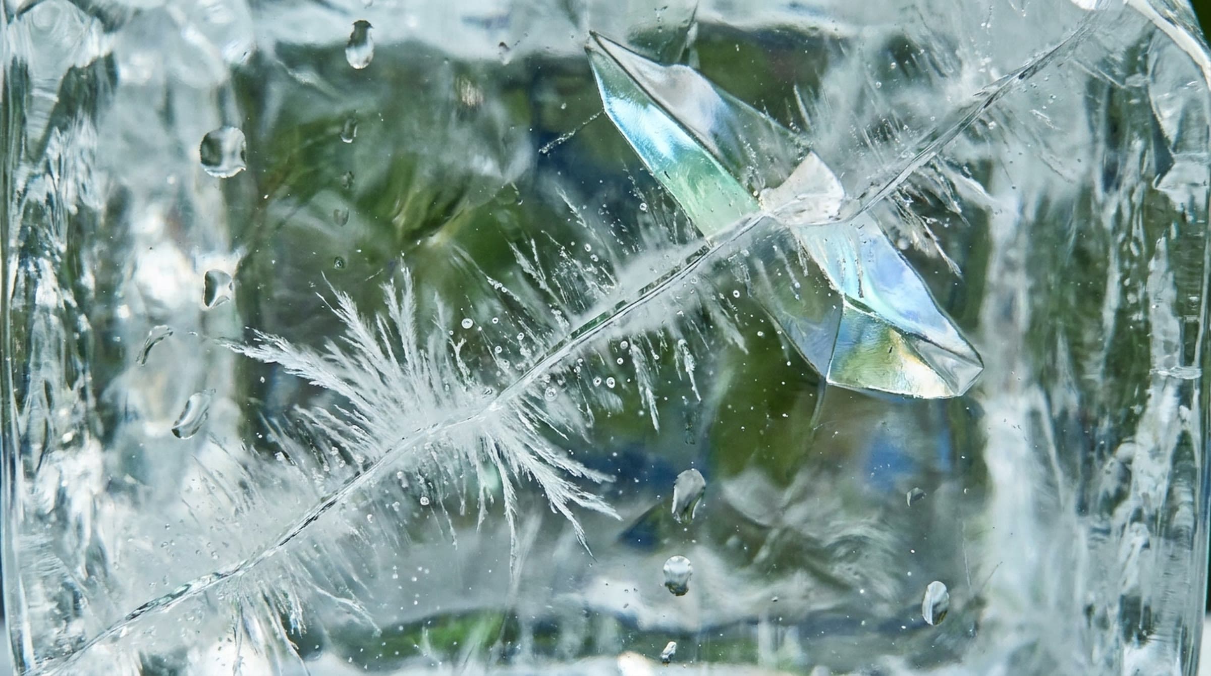 Inside a block of crystal-clear ice, a single fracture with feathered white lines and tiny bubbles, green and blue daylight showing through.
