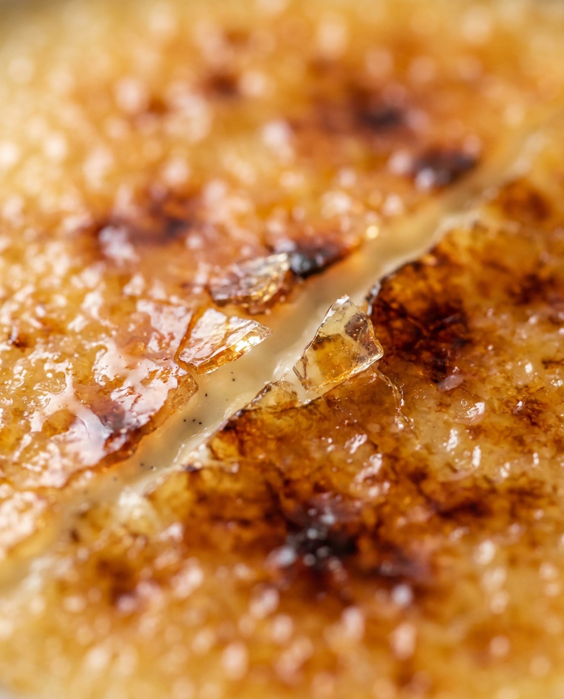 Close view of a torched caramelized sugar crust cracked open to show pale vanilla custard, amber shards lifting at the break.