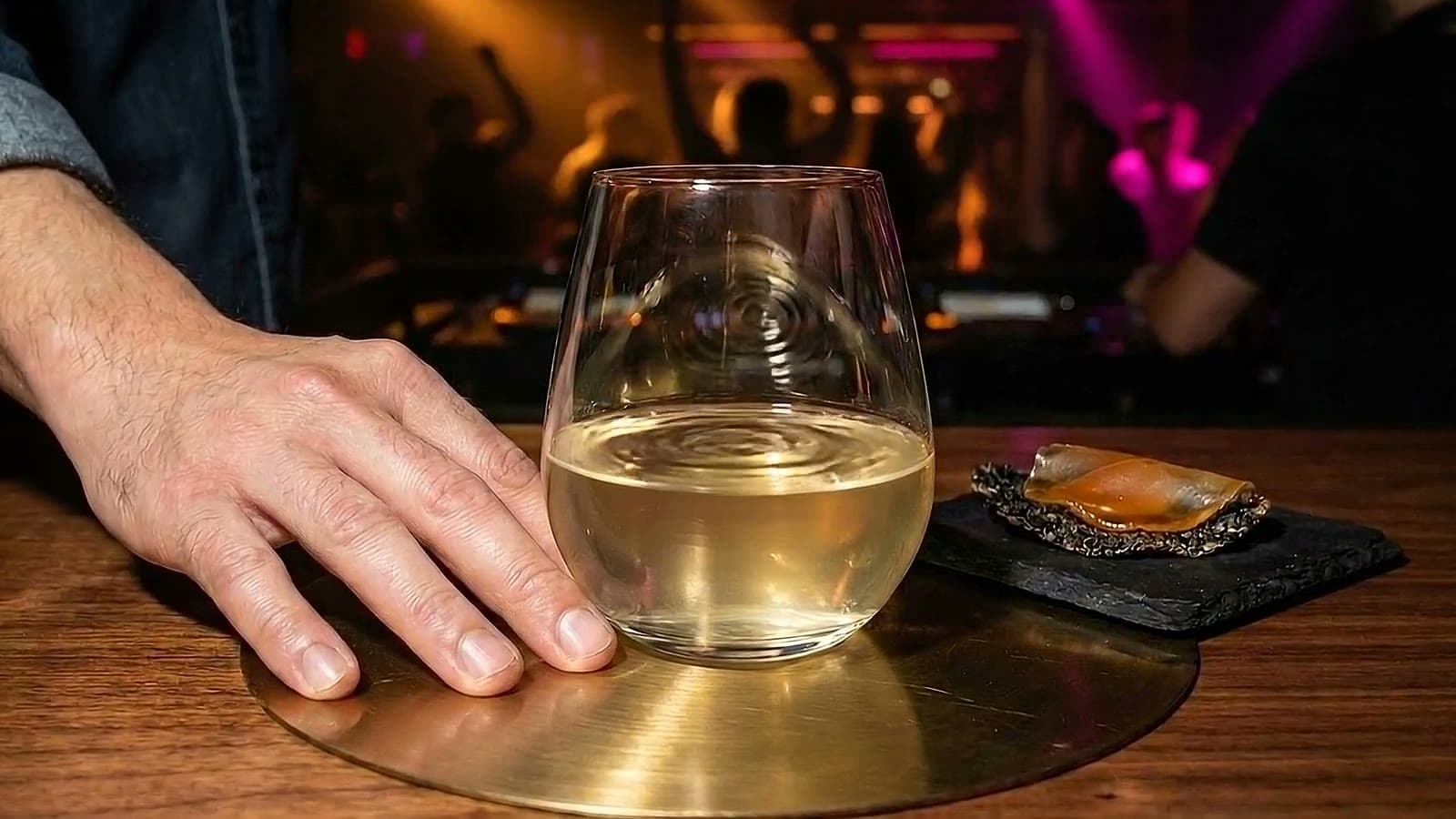 Rings ripple across the surface of a pale gold cocktail sitting on a brass plate on a nightclub bar as dancers move in the blurred background.