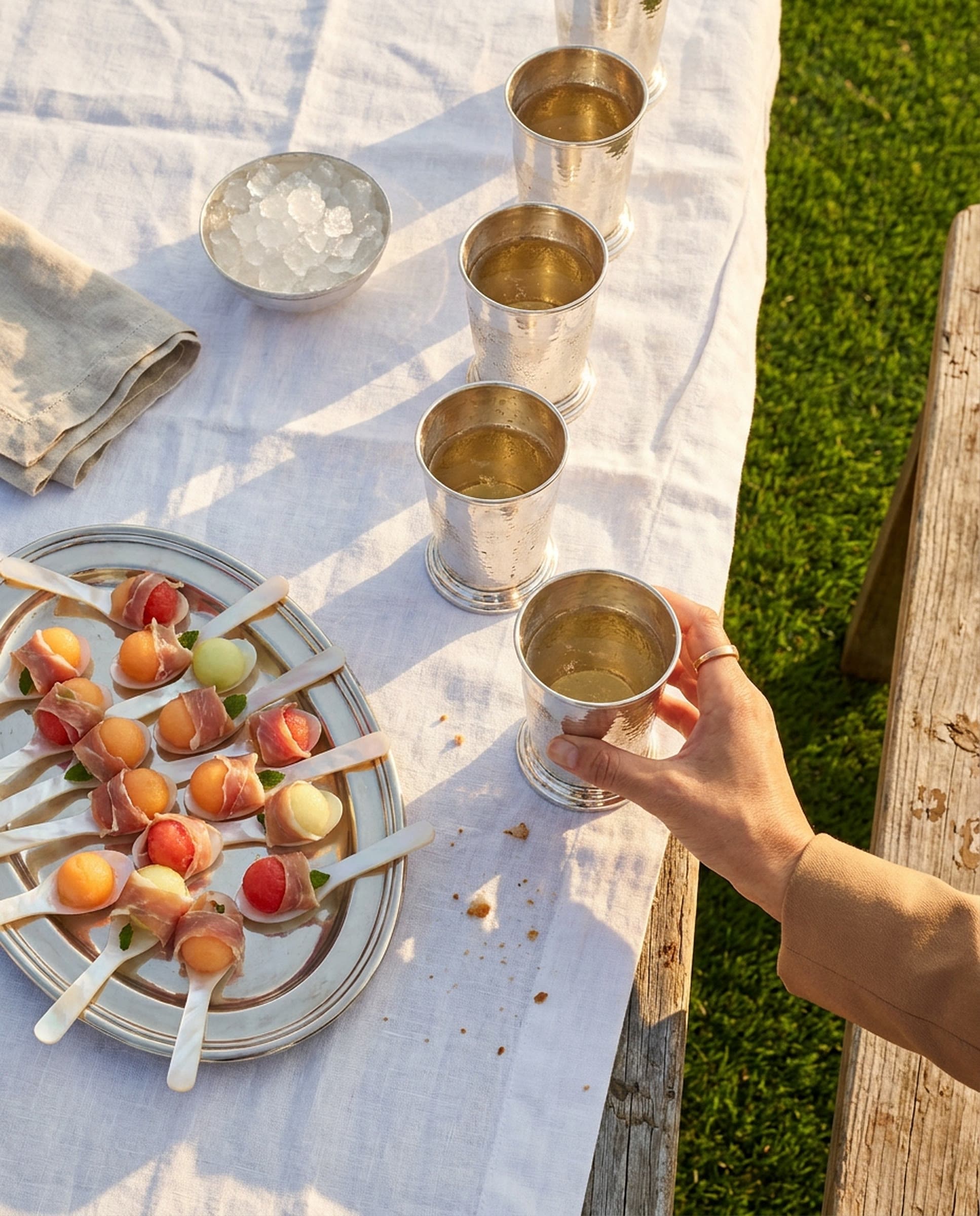 A white linen cloth on a weathered wooden table beside green polo turf in low golden sun, with a row of hammered silver cups of a pale gold drink, an oval silver tray of mother-of-pearl spoons holding melon balls wrapped in shaved ham with mint, a bowl of crushed ice, and a woman's hand in a camel sleeve lifting one cup.