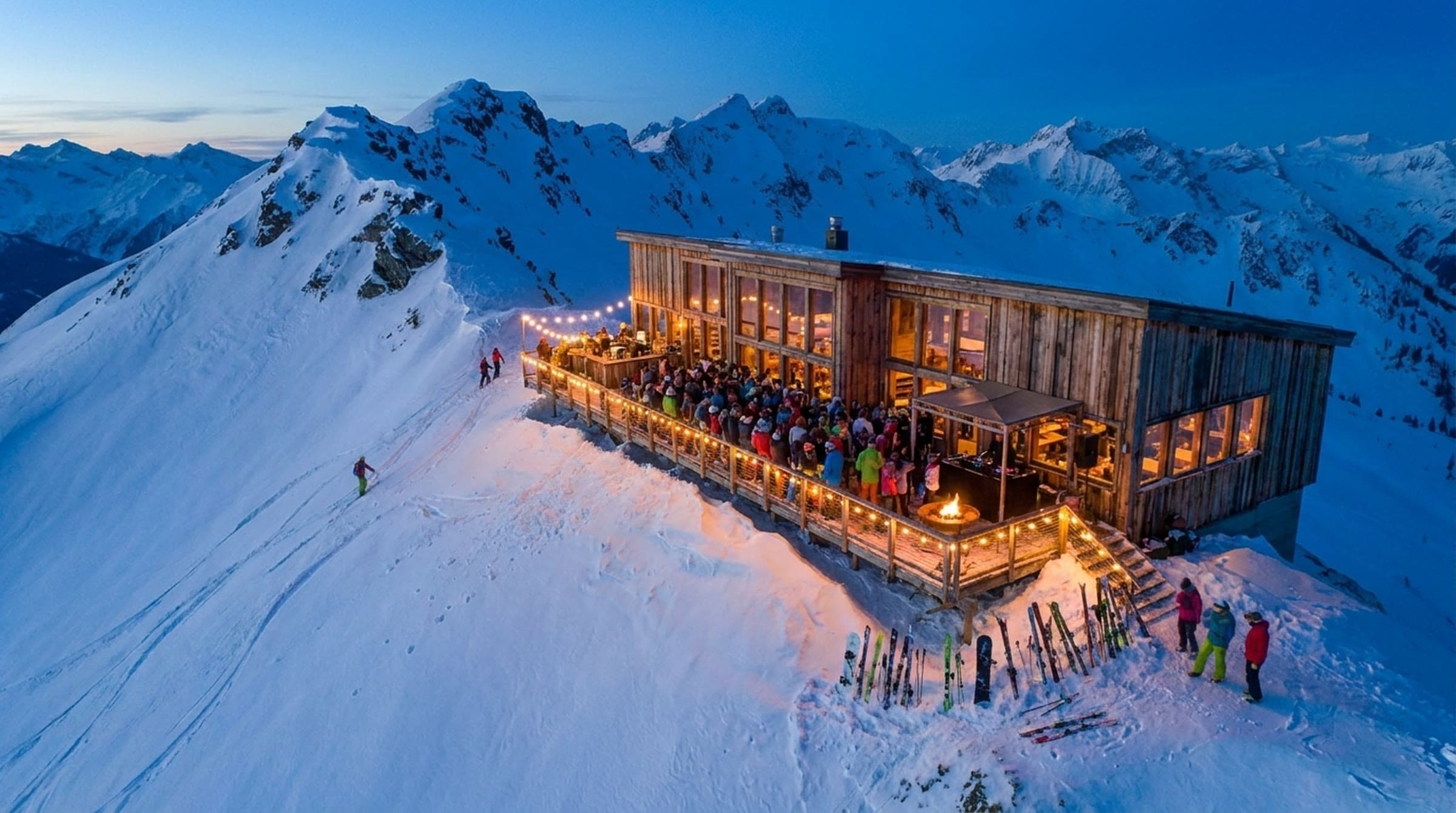 The ridge lodge at blue hour with every window lit and the full length of its deck packed for an apres party under strings of warm bulbs, skis stacked at the stairs.
