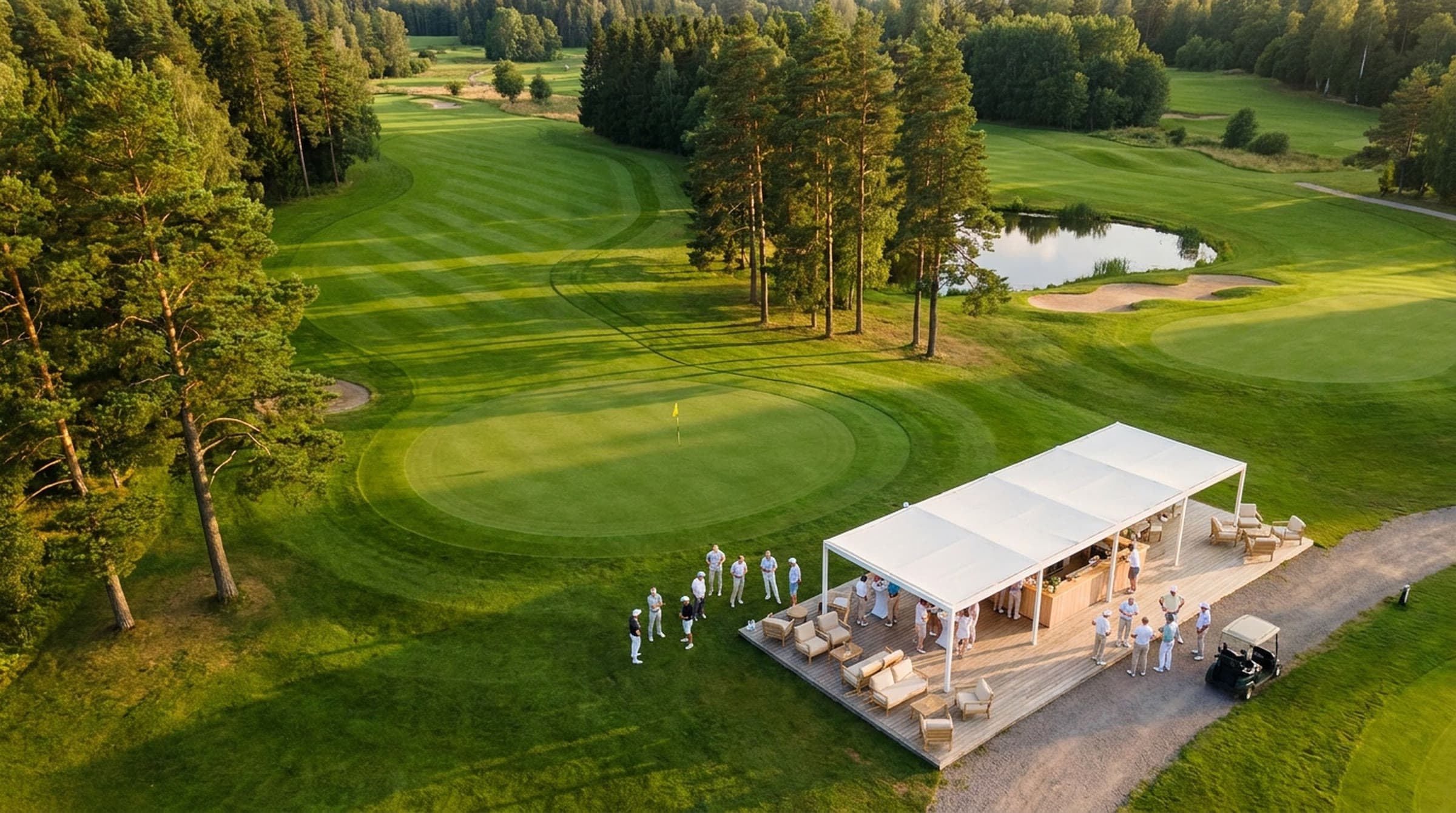 Aerial view at golden hour of a golf course's final green ringed by pines and a pond, with an open white-canopy lounge on a timber deck beside it, guests gathered at the bar and a golf cart parked on the path.