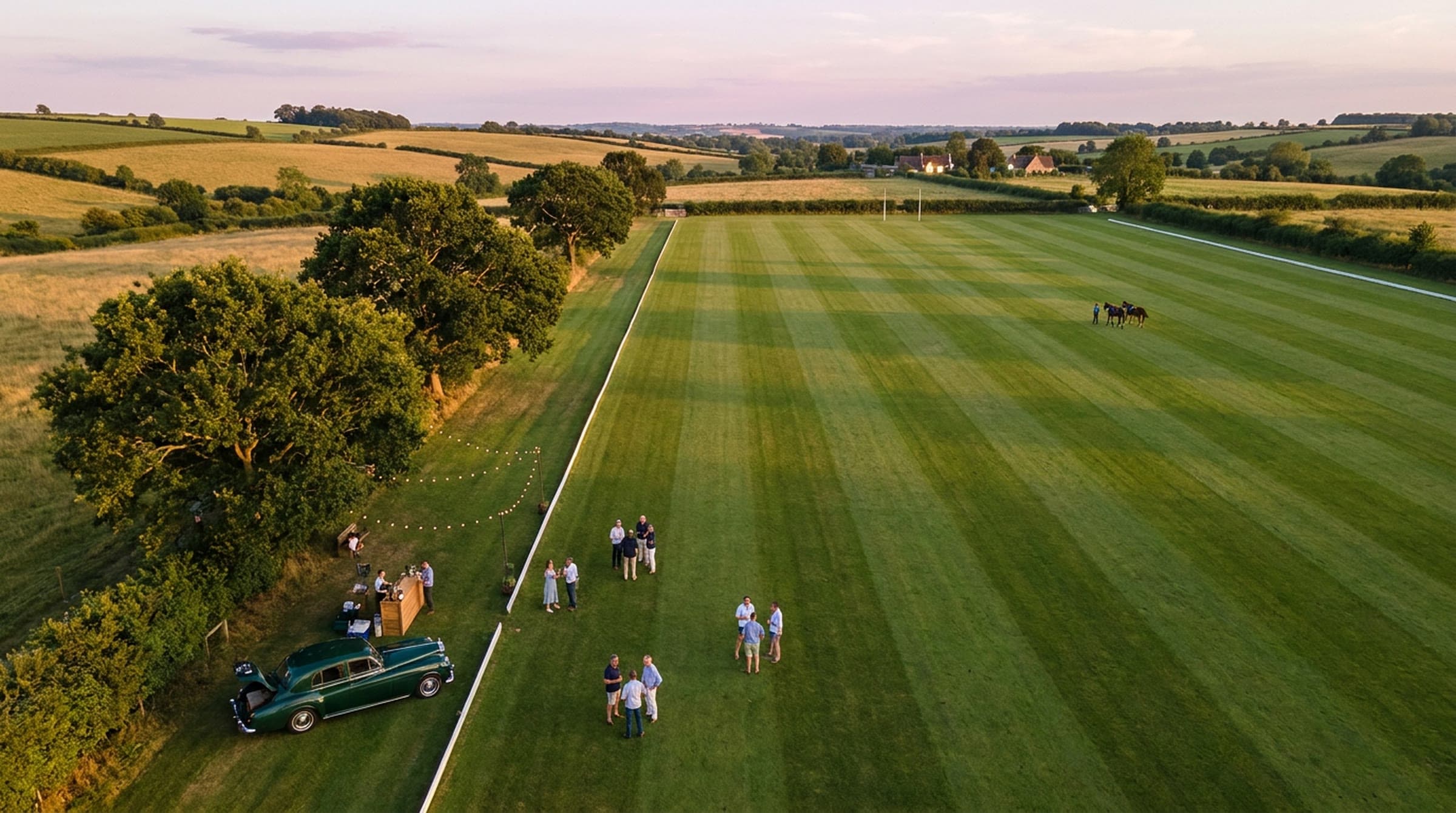 Aerial view at dusk of a striped green polo field in rolling countryside, a vintage green sedan parked under old oaks on the sideline beside a small bar and string lights, small groups of guests on the grass and two horses at the far end.