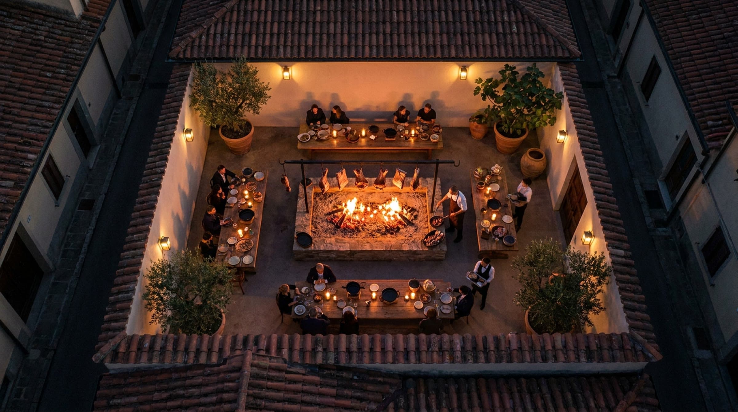 Top-down view at night into a walled courtyard with a glowing ember fire pit under a bar of hanging meats, a cook at the fire and guests at candlelit wooden tables, surrounded by olive trees and terracotta rooftops.