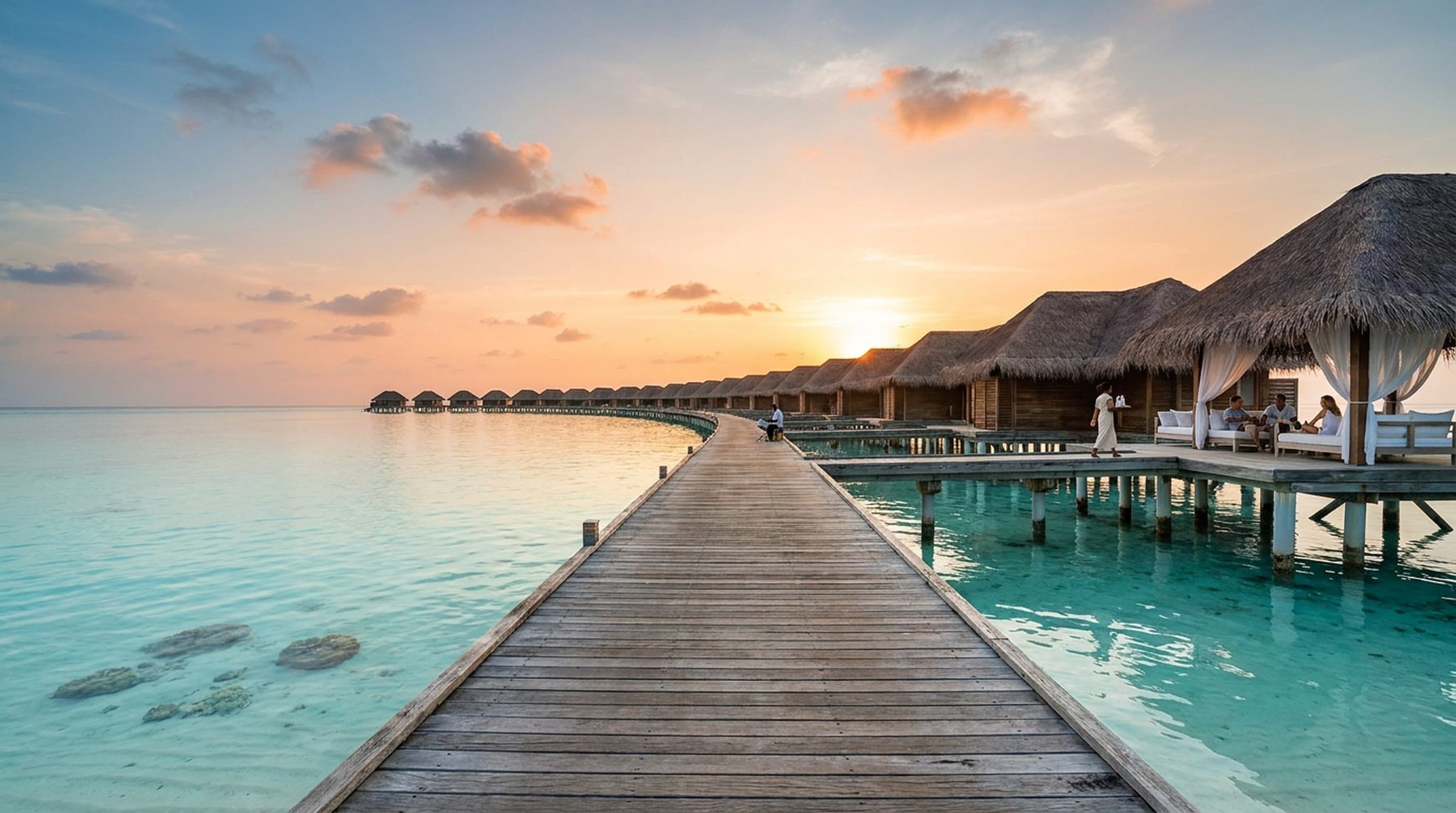A long wooden boardwalk stretching over a clear turquoise lagoon at sunset toward a curving row of thatched over-water cabanas, a server carrying an ice tray toward friends relaxing on white daybeds under sheer curtains.
