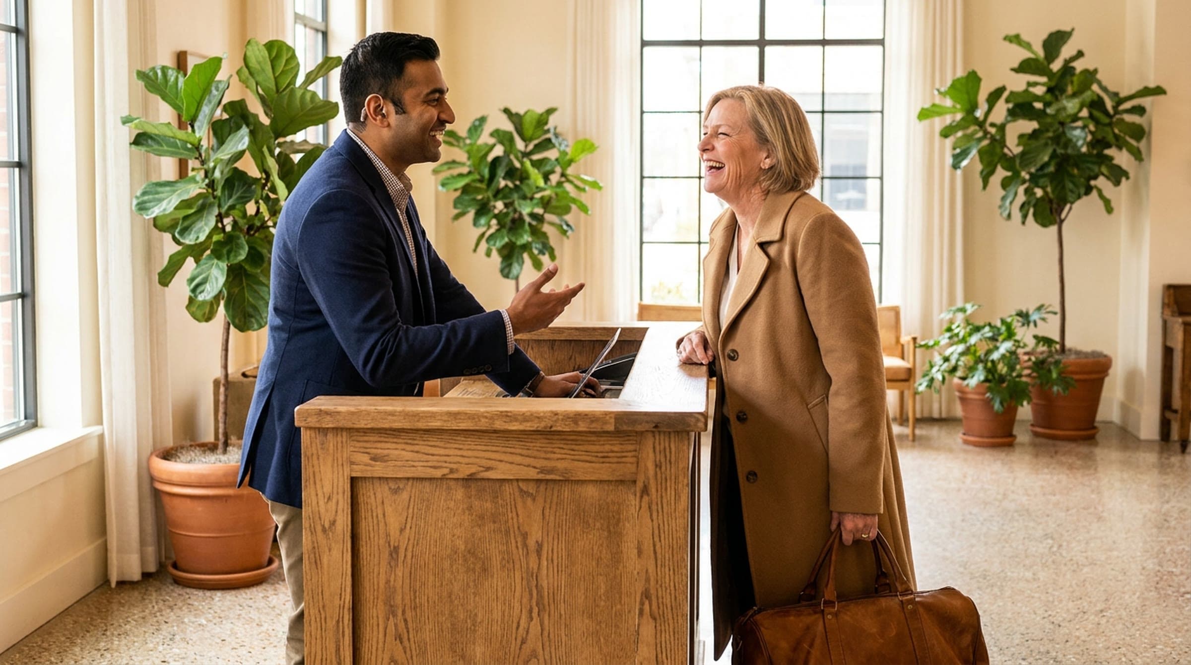 A front desk associate in a navy jacket laughs with an arriving guest in a camel coat across an oak desk in a bright lobby filled with plants.