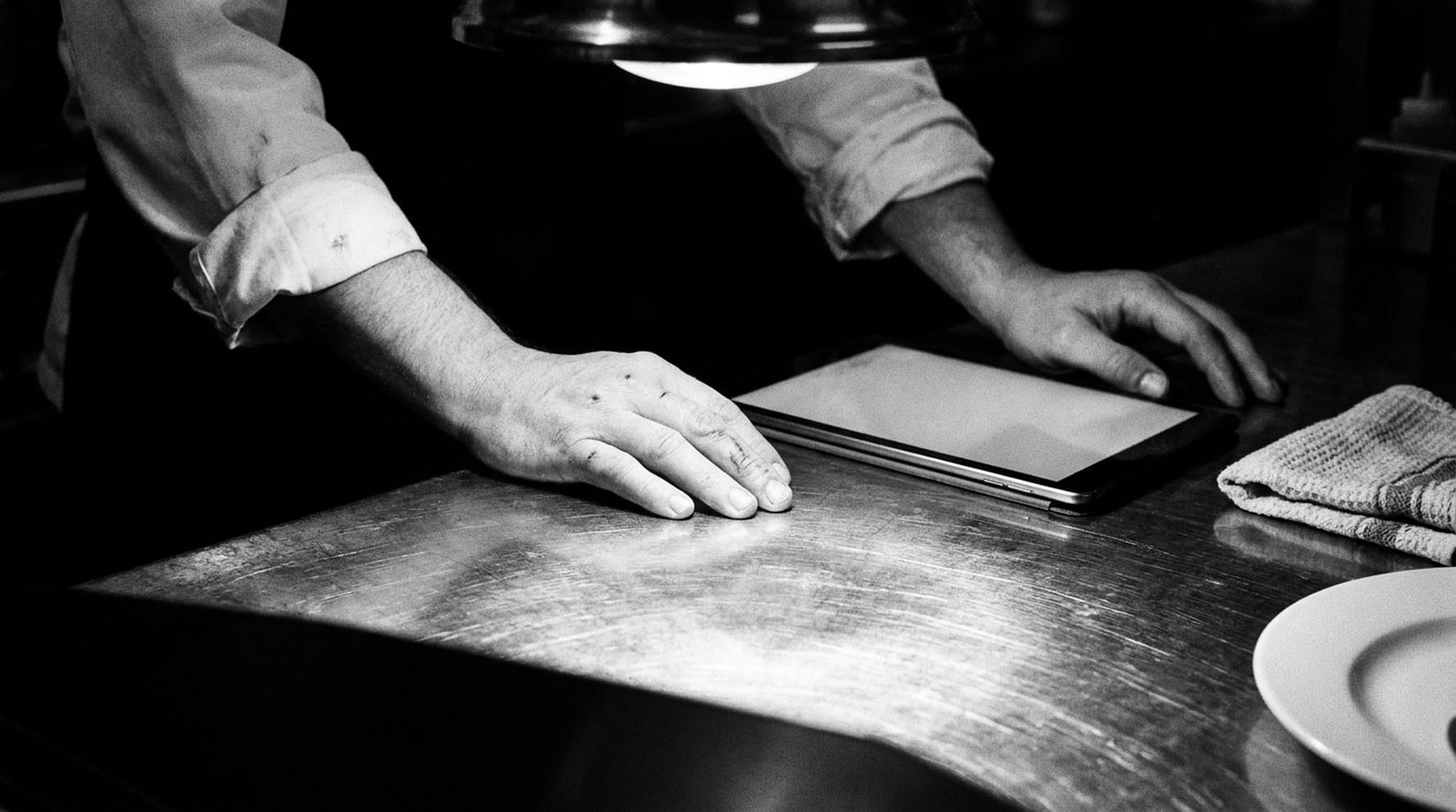 A cook's hands rest on a steel kitchen pass under a single hard overhead lamp, one hand flat on the steel and the other beside a tablet lying screen up, with a folded towel and the edge of a white plate nearby, in black and white.