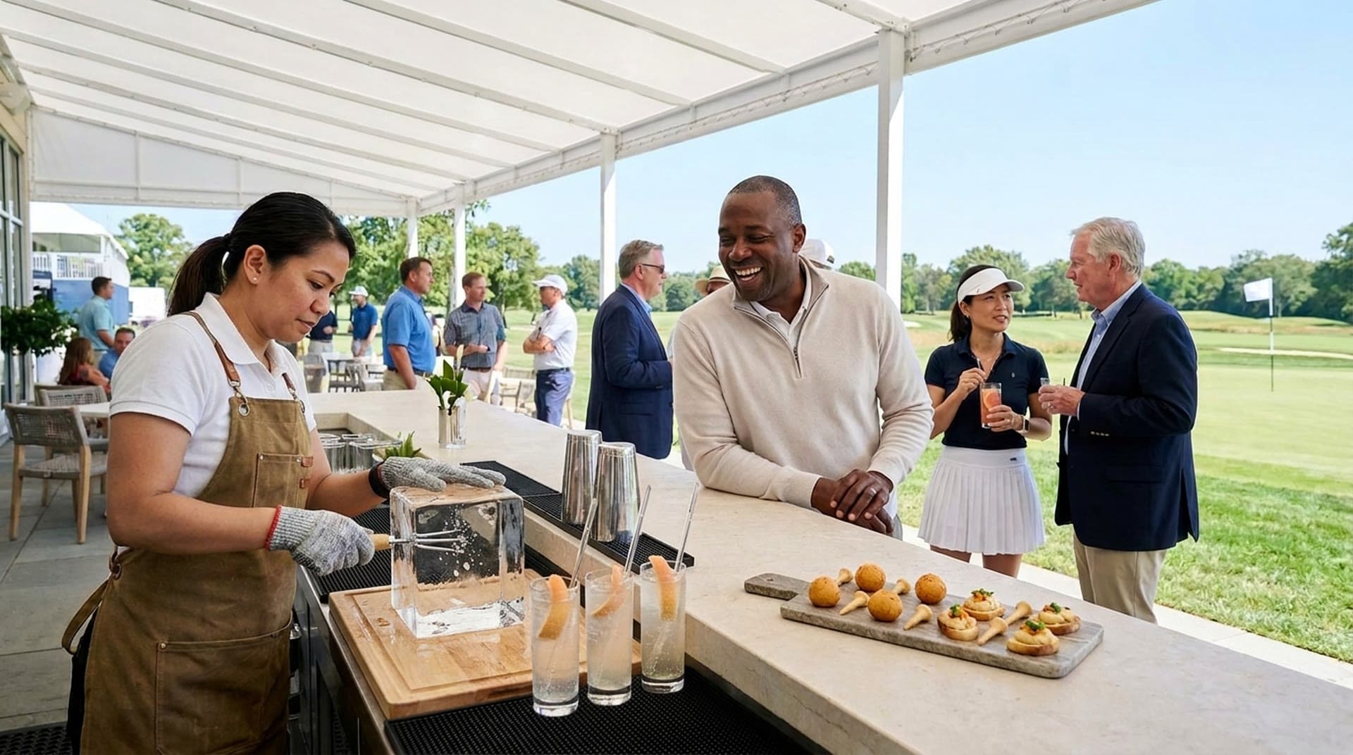 A bartender in a canvas apron and cut-resistant gloves works a large block of clear ice on a wooden board at an outdoor bar under a white canopy, while guests in golf attire laugh and talk across the bar with a fairway and a flag on the green behind them.