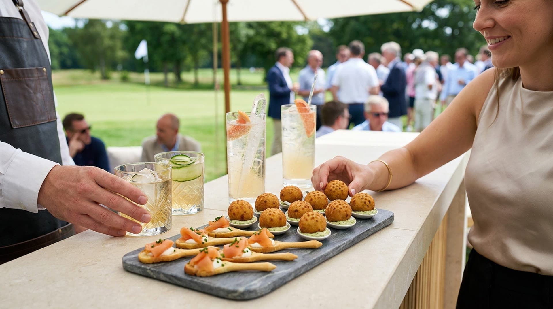 A stone board of round golden croquettes shaped like golf balls on small spoons of herb sauce and tee-shaped crostini topped with smoked salmon, beside highballs with long ice spears and a crystal rocks glass, as a guest reaches for a croquette on a golf course terrace.