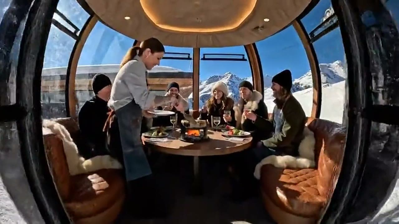 Dinner for six inside a glass gondola cabin, a server at the table and snowy peaks through the glass.